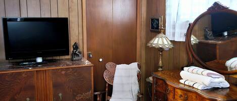 A room with a wooden wardrobe, a television, a chair with a blanket, a dresser with a mirror, and a bed.