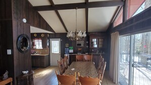 A dining area with a long wooden table and chairs, a chandelier, and a view through large windows.