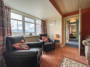 A cozy living room with two leather sofas, a small table with a lamp, and a view of the outdoors through large windows.