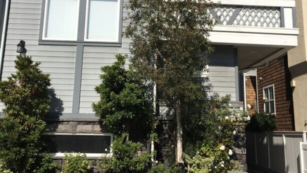 Une maison moderne à deux étages, avec une façade grise, un balcon orné d’une treille blanche et un jardin bien entretenu, agrémenté de divers arbustes et d’un arbre.