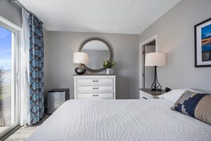 A bedroom with a large bed, a dresser with drawers, a mirror, a small table with a lamp, and a window with curtains. - First Floor Standard Suite Bay View (North East)