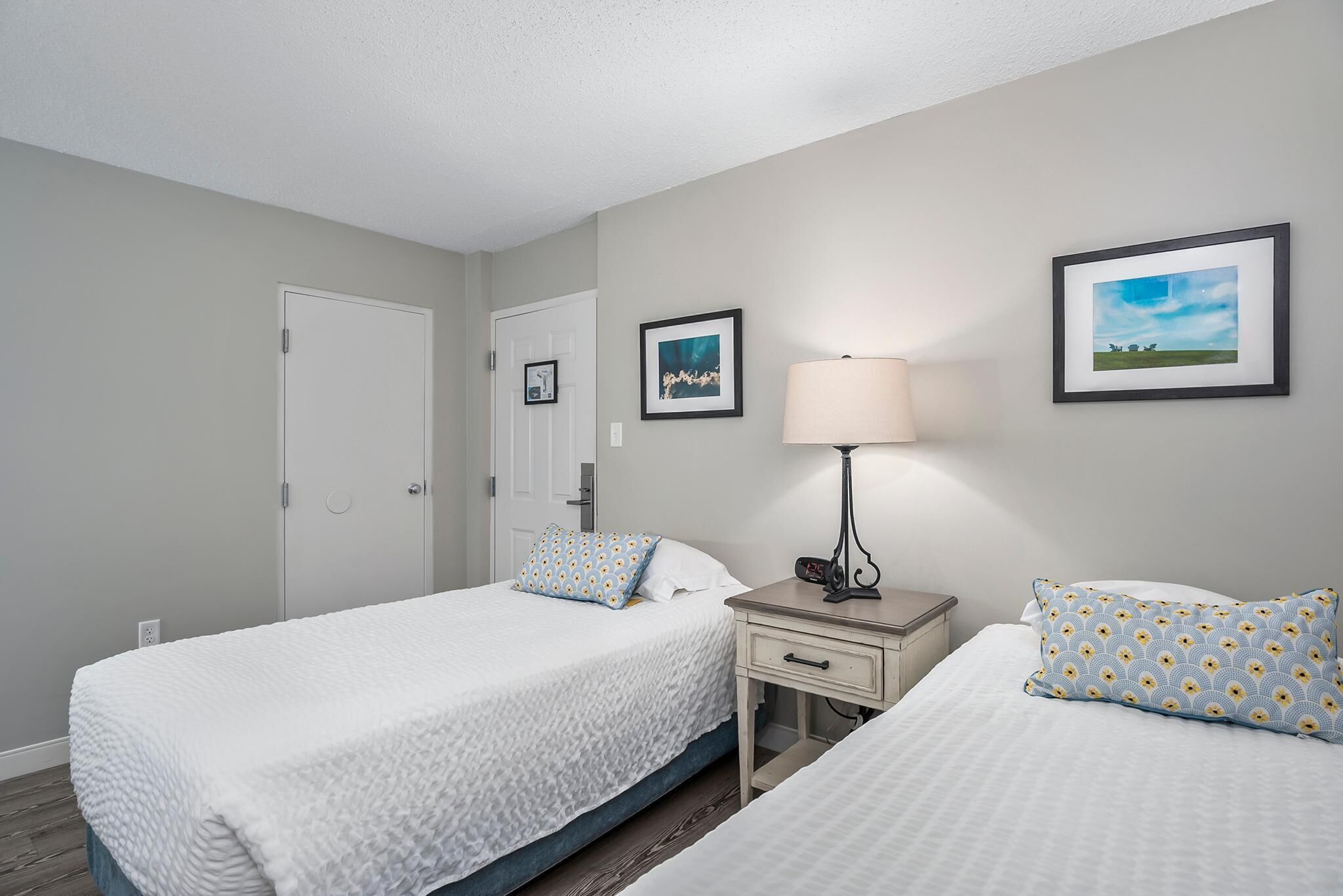 A hotel room with two beds, a bedside table with a lamp, and framed pictures on the walls.