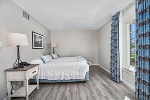 Un dormitorio con cama, mesitas de noche, lámpara, un cuadro en la pared y ventana con cortinas.