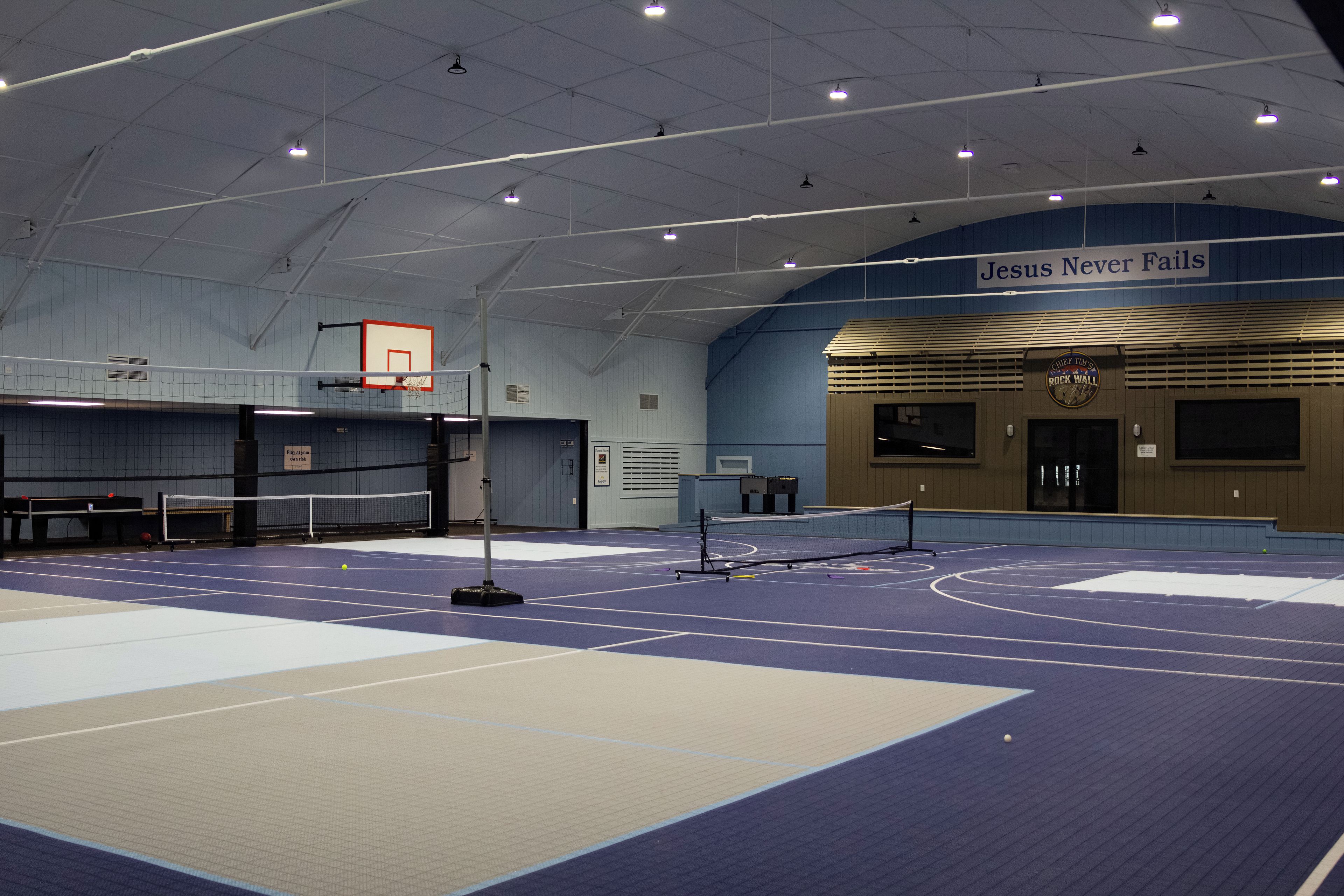 An indoor sports facility with a basketball court, a volleyball net, and a sign reading 'Jesus Never Fails'.