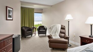 A hotel room with a brown leather chair, a small table, a lamp, and a view of the outdoors through a window with green curtains. - Fantastic Suite With King Bed and Beautiful Bay View! (North East)