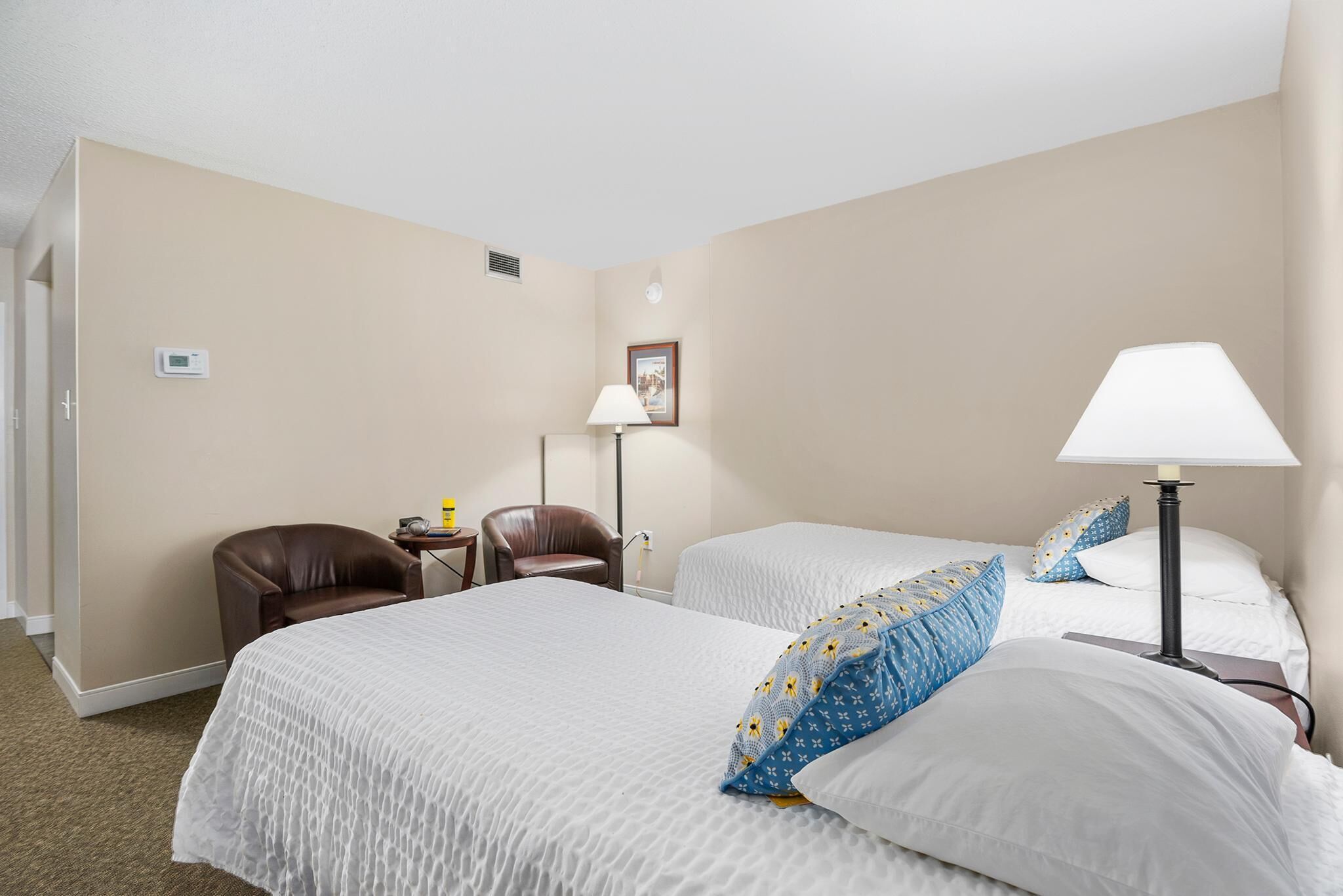 A hotel room with two beds, a desk, a chair, a lamp, and a picture on the wall.