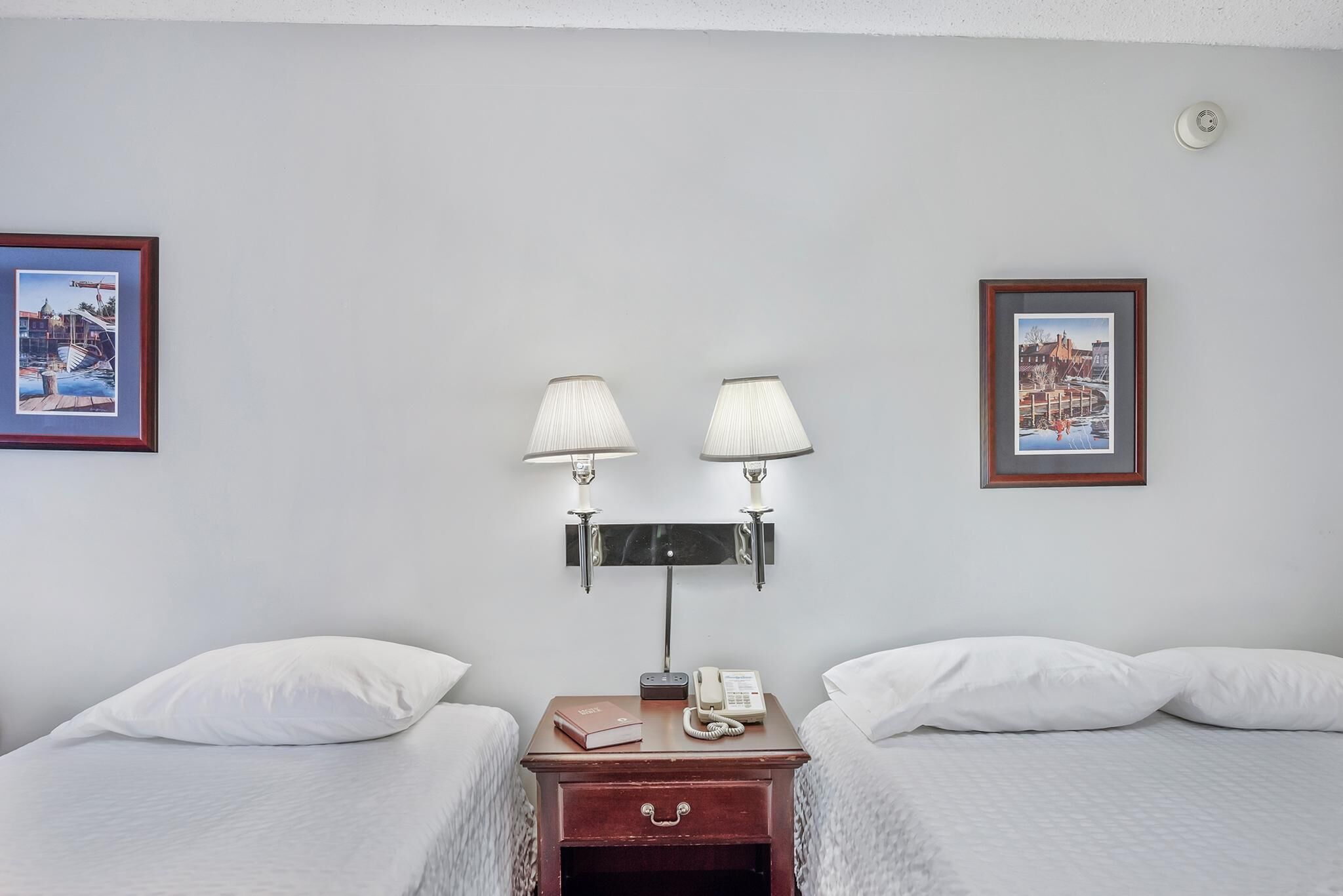 A hotel room with two beds, a nightstand with a lamp, a phone, and framed pictures on the wall.