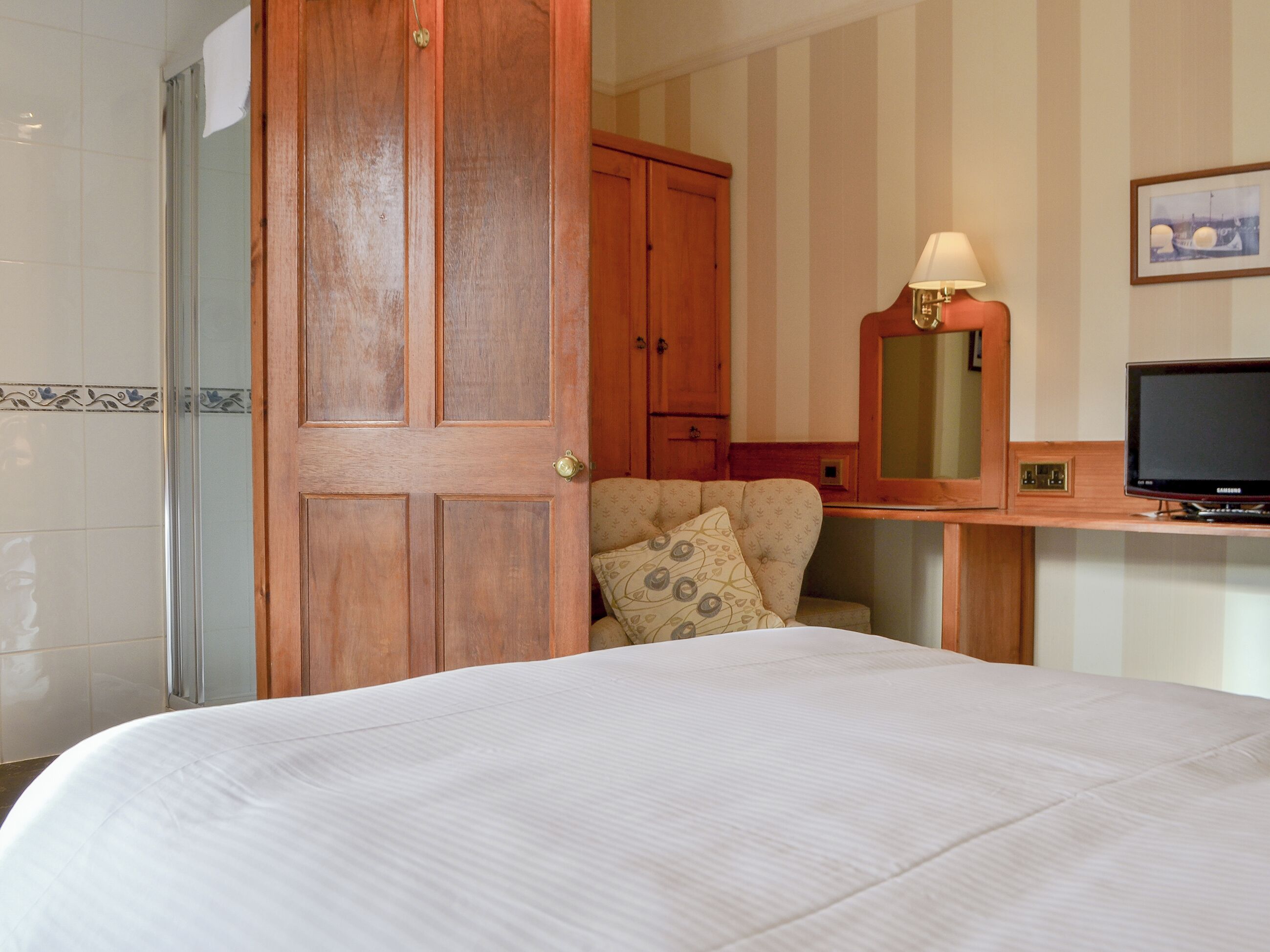 A hotel room with a bed, a wooden wardrobe, a desk with a lamp, a television, and a chair with a patterned cushion.