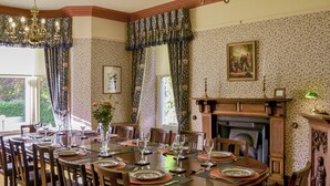 A dining room with a large round table set for six, surrounded by wooden chairs. There is a fireplace with a mantel, a chandelier, and a framed picture above it.