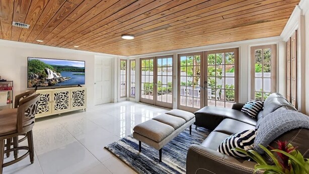 A spacious living room with a wooden ceiling, a large flat-screen TV, a sofa, a coffee table, and a dining area with a wooden table and chairs. - Seahorse Villa 1 By Dream Vacation (Fort Lauderdale)