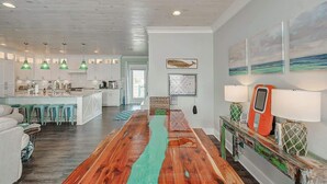 A spacious living area with a wooden floor, a kitchen in the background, and a dining table with a colorful runner. - The Oceanic, Beachfront Oasis*Sleeps 20*Hot Tub (Crystal Beach)