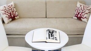 A beige sofa with red tree-patterned pillows, a white coffee table, and an open book with a black and white illustration. - King Room with Balcony (Sorrento)