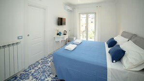A bedroom with a bed, a small bedside table, a TV, a radiator, and a window with curtains. - King Room with Balcony (Sorrento)