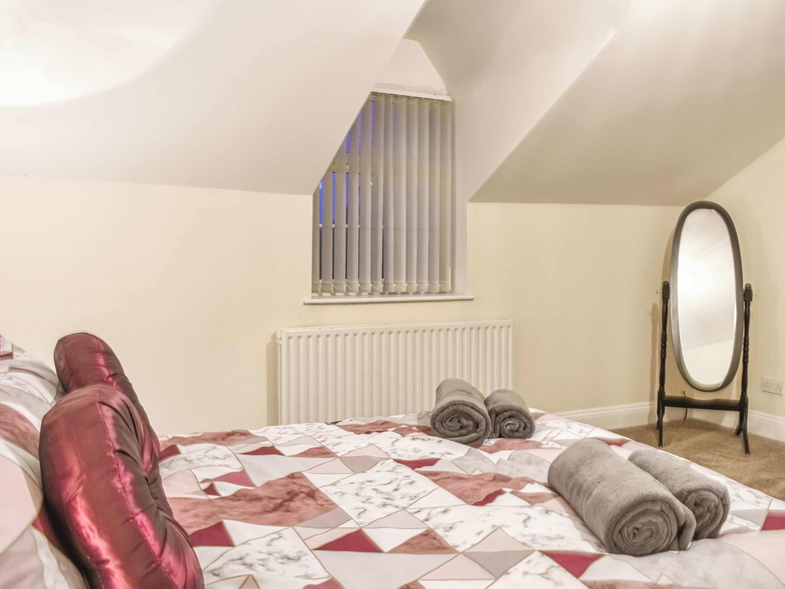 A bedroom with a bed, a radiator, a window with blinds, a mirror, and towels.