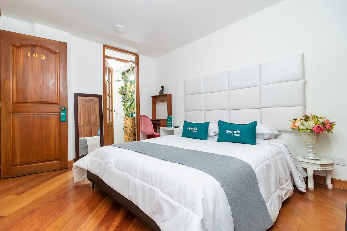 A hotel room with a large bed, a wooden door with the number 103, a small table with a vase of flowers, and a chair.