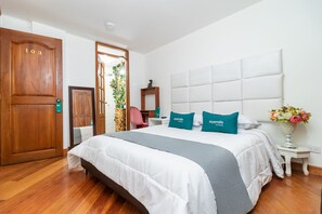 A hotel room with a large bed, a wooden door with the number 103, a small table with a vase of flowers, and a chair. - Ayenda Dorado Boutique (Bogotá)