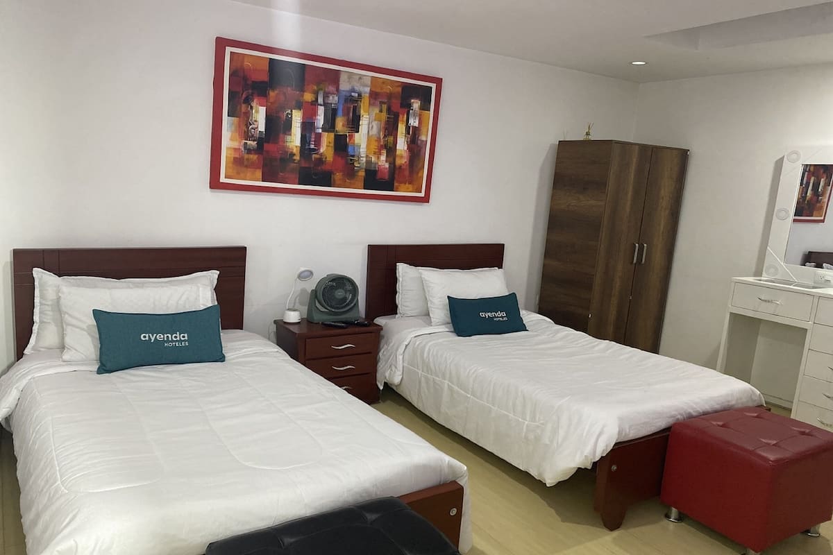 A hotel room with two beds, each with white bedding and teal pillows, a wooden wardrobe, a vanity with a mirror, and a red ottoman.