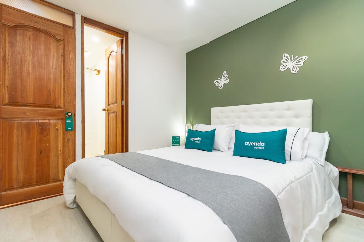 A hotel room with a large wooden door, a white bed with teal pillows, and a green wall with butterfly decorations.