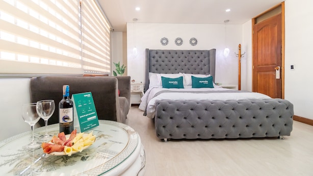 Una habitación de hotel con una cama, un banco tapizado en gris, una mesa de vidrio con una botella de vino y un plato de comida, y una ventana con persianas.