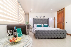 Una habitación de hotel con una cama, un banco tapizado en gris, una mesa de vidrio con una botella de vino y un plato de comida, y una ventana con persianas.