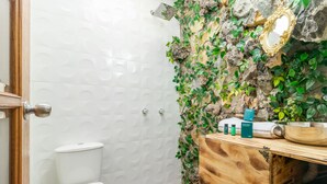 Un baño con inodoro blanco, tocador de madera, espejo y zona de ducha con pared de piedra y plantas.