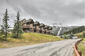A row of log cabin-style lodges with a mountain backdrop, a winding road, and a truck parked nearby. - Mountain Views + Hot Tub: Big Sky Resort Condo! (Big Sky)