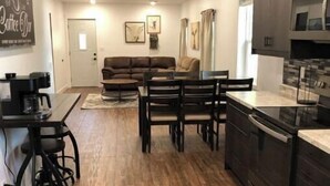 A modern kitchen with dark cabinetry, a dining area with a wooden table and chairs, and a living room with a brown sofa.
