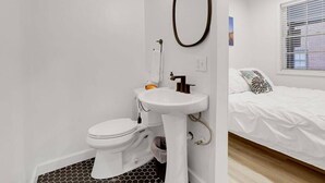 A bathroom with a toilet, sink, and mirror. There is a bed with a patterned pillow in the adjacent room.