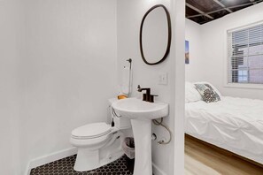 A bathroom with a toilet, sink, and mirror. There is a bed with a patterned pillow in the adjacent room. - Minimalist Modern Suite Near German Village (Columbus)