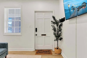 A clean, modern entryway with a white door, a potted plant, a doormat, and a flat-screen TV on the wall.