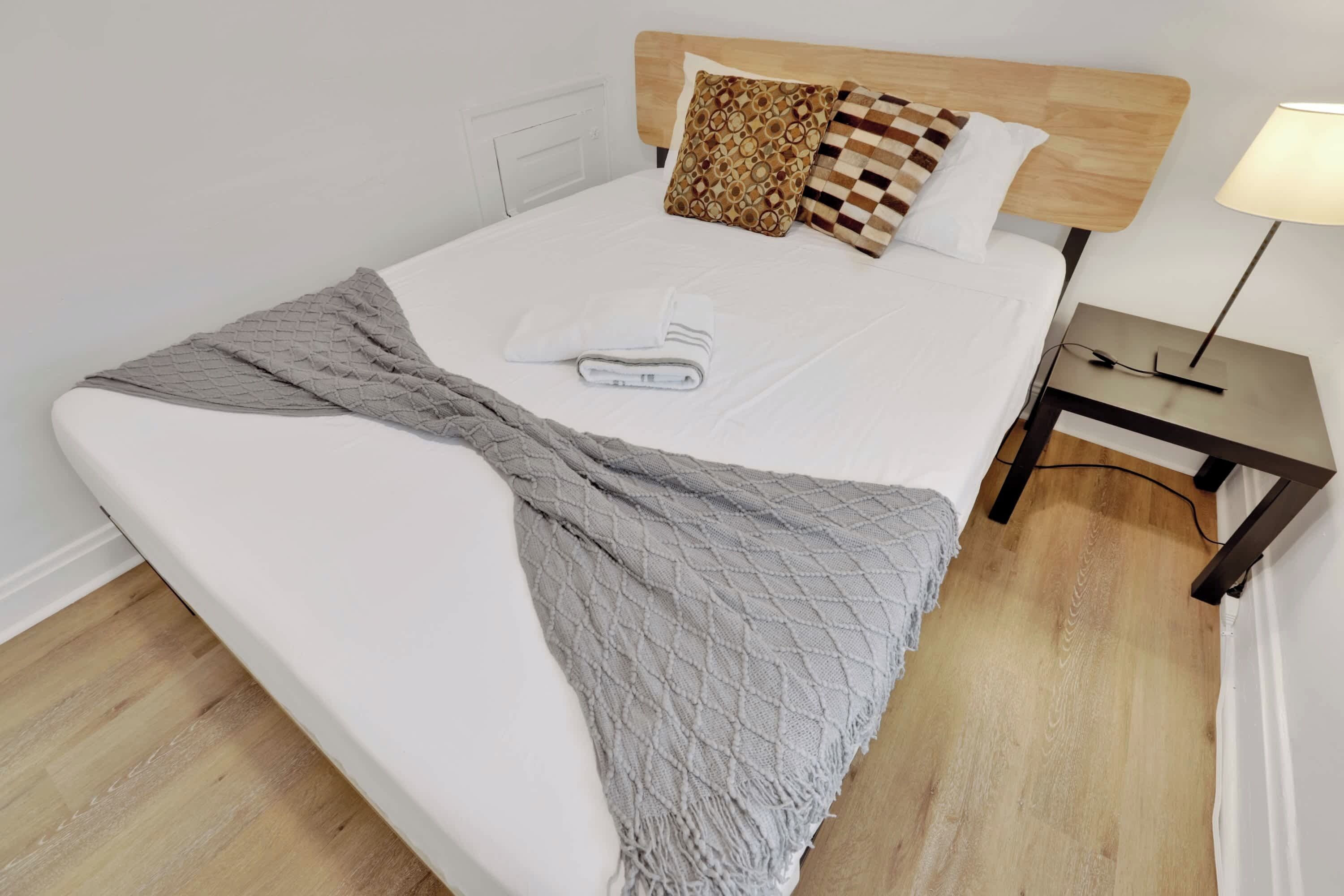 A neatly made bed with a grey blanket, a wooden headboard, patterned pillows, and a bedside table with a lamp.