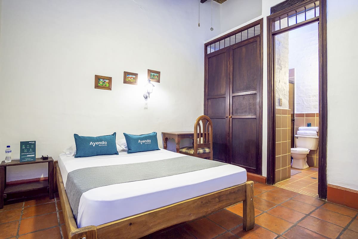 Habitación de hotel con cama, mesita de noche, silla de madera, armario y baño visible a través de una puerta abierta.