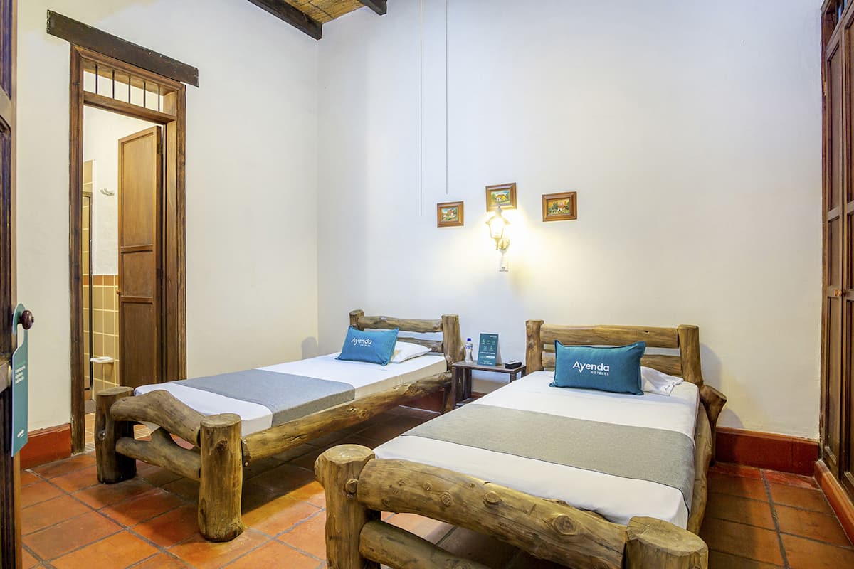 Una habitación con dos camas de madera, cada una con un almohadón que dice "Ayenda". Hay tres cuadros enmarcados en la pared, una mesita de noche con una lámpara y una puerta de madera.
