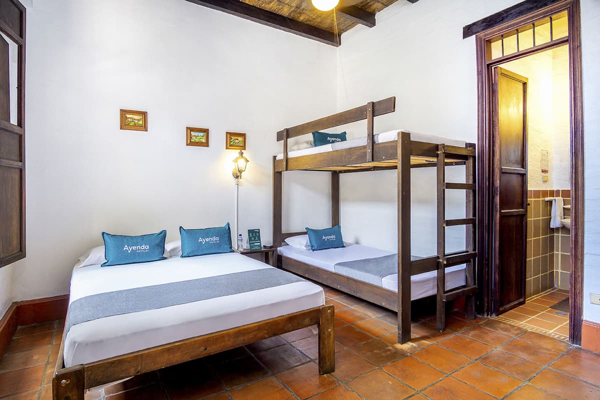 Una habitación de hotel con una cama nido de madera, una cama individual con colcha gris y una lámpara fijada a la pared.