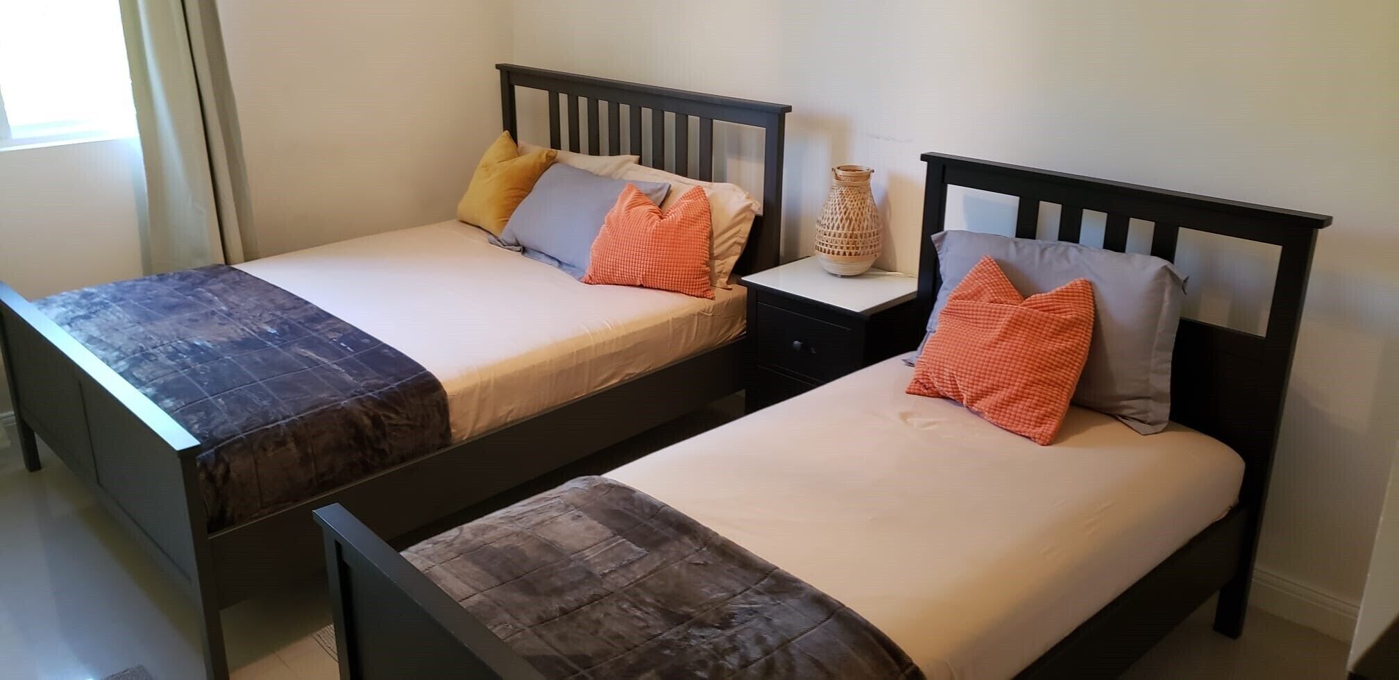 Two twin beds with wooden frames and beige bedding, a small bedside table with a vase, and a window with curtains.