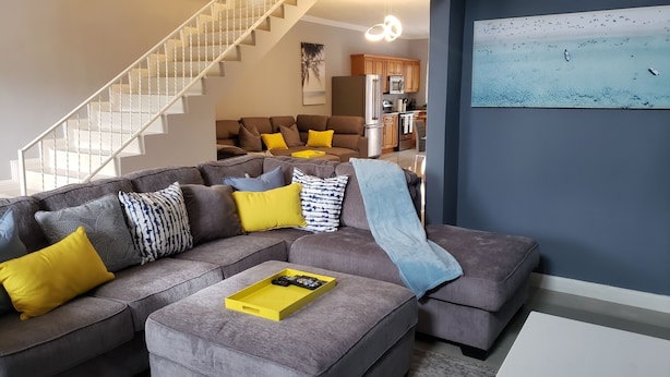 Ein modernes Wohnzimmer mit einem grauen Ecksofa, gelb-gemusterten Kissen, einem gelben Tablett und einer Treppe, die zu einer oberen Ebene führt.