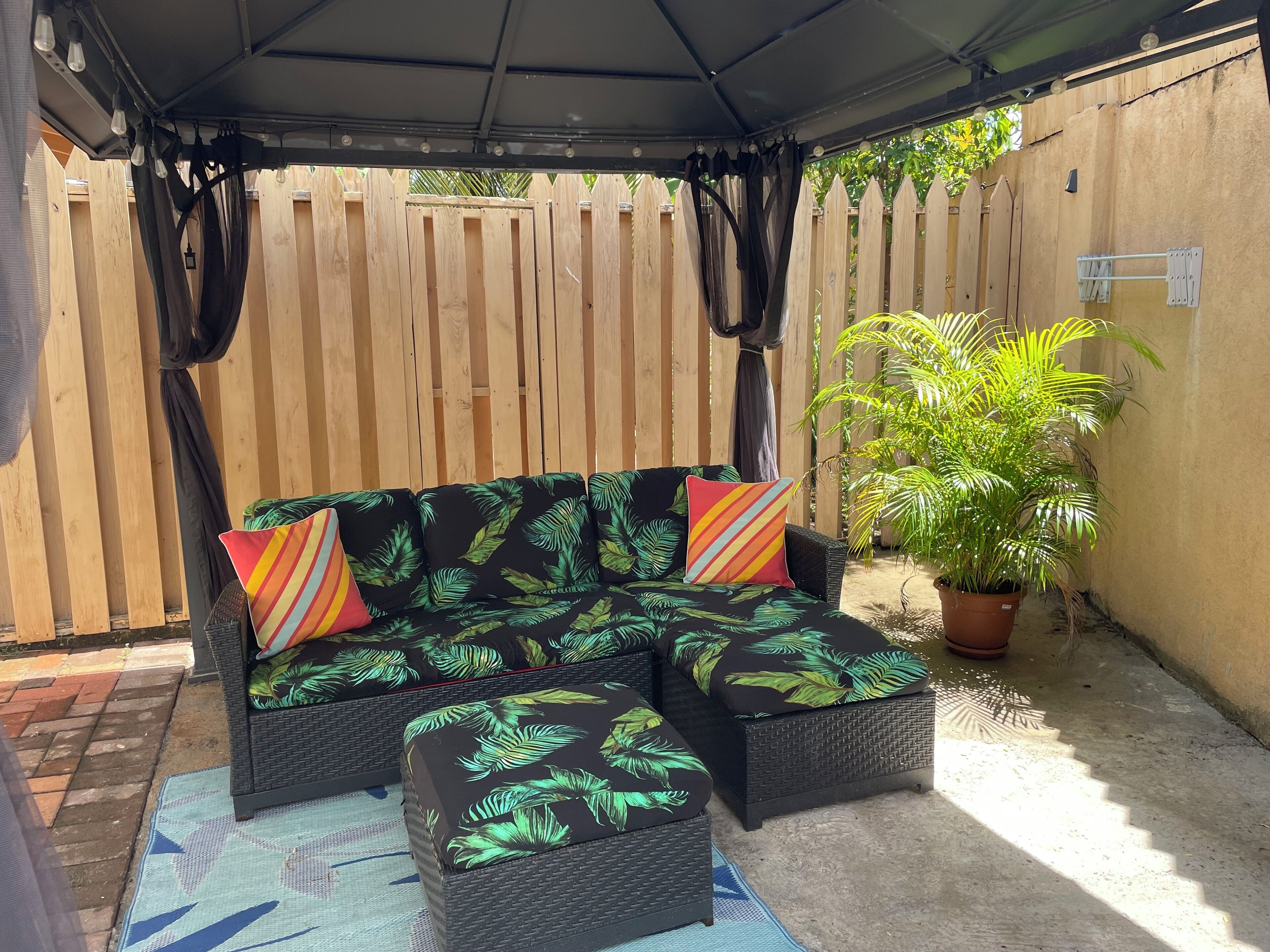 Un espace extérieur aménagé avec un salon de jardin couvert, comprenant un canapé et un pouf en osier, une plante en pot et une clôture en bois.