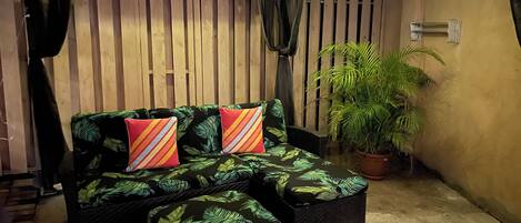 A covered outdoor seating area with a wicker sofa and ottoman, a potted plant, and a wooden fence.