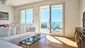 A modern living room with a sofa, a glass coffee table, and a TV. There are large sliding glass doors leading to a balcony with a view of the sea.