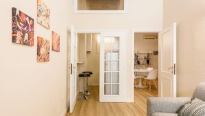 Un salon avec un canapé gris, un coin repas avec une table et des chaises blanches, et un couloir avec un plancher en bois et des œuvres d’art accrochées aux murs.