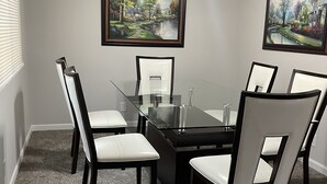 Une salle de conférence avec une table en verre, quatre chaises, deux tableaux, un ventilateur de plafond et une fenêtre avec des stores.