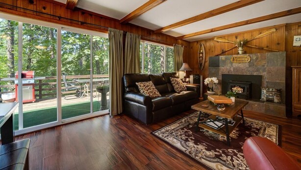 A cozy living room with a fireplace, a leather sofa, and a large window overlooking a deck. - Cabin in the Pines, 3BR, Arcade Room, Wraparound Deck, Walk to Town, Dogs OK (Twain Harte)