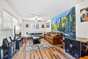 Una sala de estar con mesa de billar, un sofá de cuero marrón, un cuadro grande de un bosque y piso de madera.