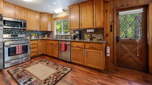 A kitchen with wooden cabinets, stainless steel appliances, a patterned rug, and a view of trees through a window. - Cabin in the Pines, 3BR, Arcade Room, Wraparound Deck, Walk to Town, Dogs OK (Twain Harte)
