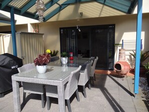Un patio al aire libre con una mesa de vidrio rodeada de sillas de mimbre, una parrilla negra y plantas en macetas.