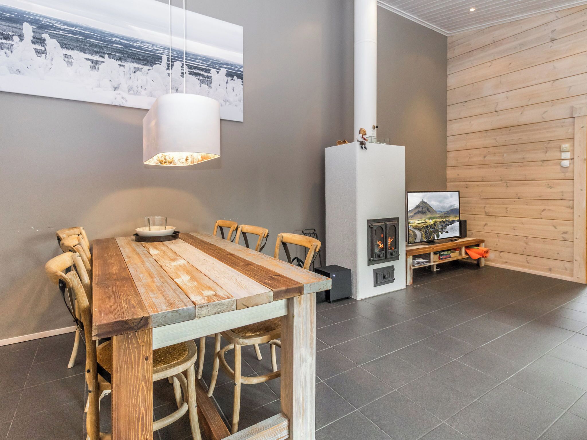A modern dining area with a wooden table and chairs, a hanging light fixture, a fireplace, and a large window with a view of a snowy landscape.