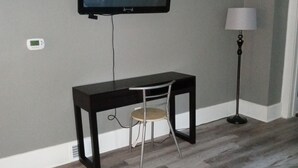 A living room with a flat-screen TV mounted on the wall, a small table with a chair, a floor lamp, and a glass coffee table.