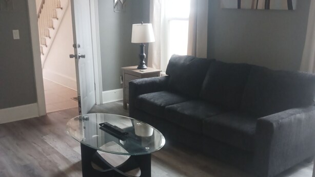 A living room with a glass coffee table, a dark sofa, a lamp on a side table, and a staircase in the background.