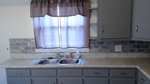 A kitchen with grey cabinets, a double sink, and a window with a brown valance.