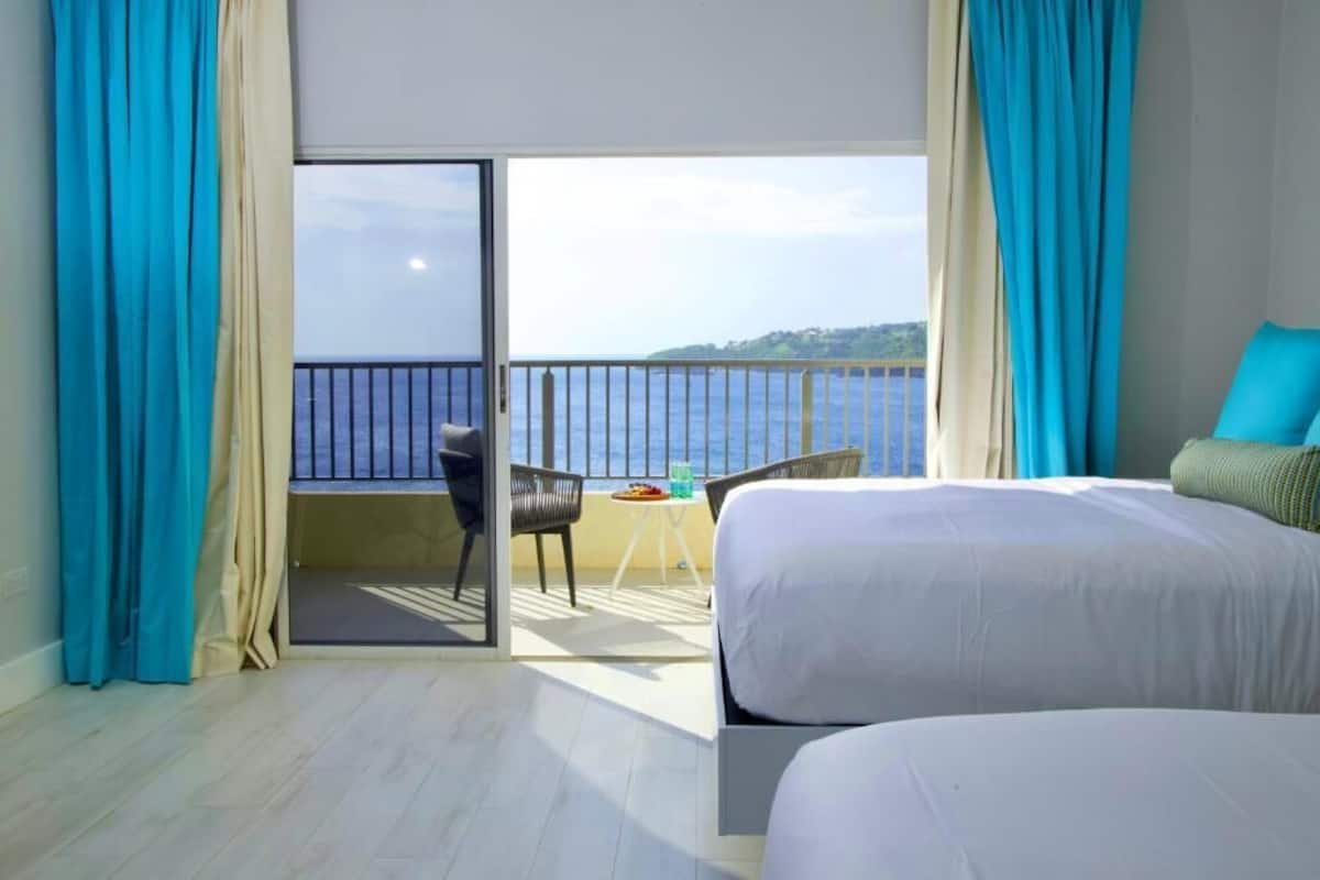 Habitación de hotel con dos camas, un balcón y vistas al mar, además de una mesa con sillas.